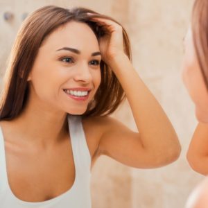 Beautiful young woman touching her hair with hand and smiling while standing in front of the mirror