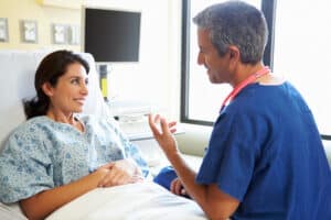 Female patient in hospital gown smiling and speaking with a male doctor, discussing anesthesia options before cosmetic surgery