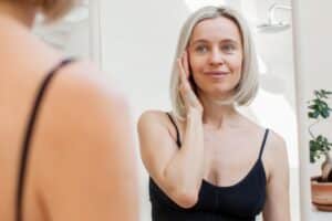 Woman looking in the mirror and pulling up part of her face to visualize a mini facelift